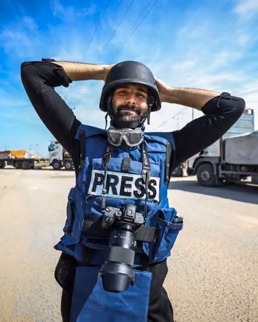Motaz Azaiza in a press vest and helmet, a camera at his chest, with trucks on the road behind him